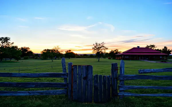 A serene ranch scene in 4K Ultra HD showcases a wooden fence gate with a lush green pasture and a charming red-roofed building against a vibrant sunset sky.