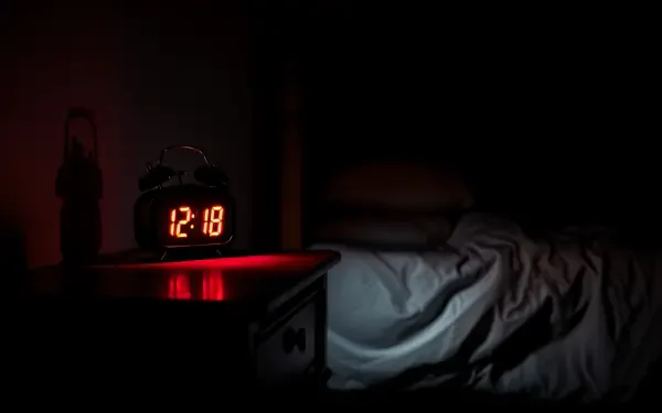 A dark scene featuring a red-lit alarm clock displaying 12:18, placed on a bedside table next to rumpled white bedding, all captured in crisp 4K Ultra HD quality.