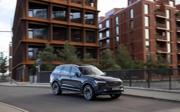 A sleek Volvo XC90 Facelift drives along a modern urban street, showcasing its sophisticated design against a backdrop of contemporary architecture. 4K Ultra HD quality.
