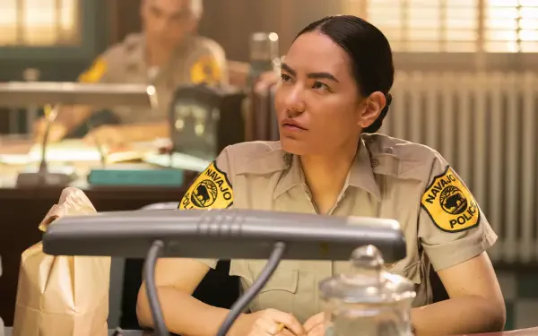 Jessica Matten portrays a police officer in a scene from the TV show Dark Winds. The image captures a moment of focus in a detailed office setting. HD desktop wallpaper reference.