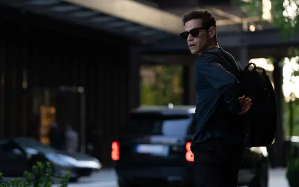 Rami Malek stars in The Amateur (2025), captured in this striking 8K Ultra HD wallpaper. The image shows him looking back while wearing sunglasses and a backpack.