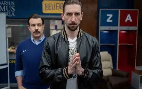 A scene from the TV show Ted Lasso featuring Jason Sudeikis and Maximilian Osinski, captured in an 8K Ultra HD wallpaper. The characters appear in a thoughtful pose against a vibrant backdrop.