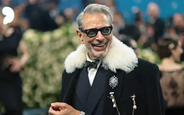 Jeff Goldblum smiling at the Met Gala, dressed in a stylish black coat with a white fur collar, captured in 4K Ultra HD as a desktop wallpaper and background.