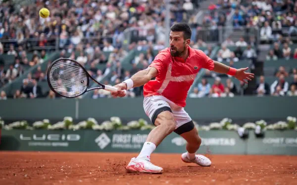 HD desktop wallpaper capturing Novak Djokovic in action during a high-intensity tennis match on a clay court, with a packed stadium in the background.