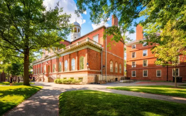 Sunny view of a historic Harvard University building in Cambridge, surrounded by lush green park trees and pathways, captured in HD for a desktop wallpaper background.