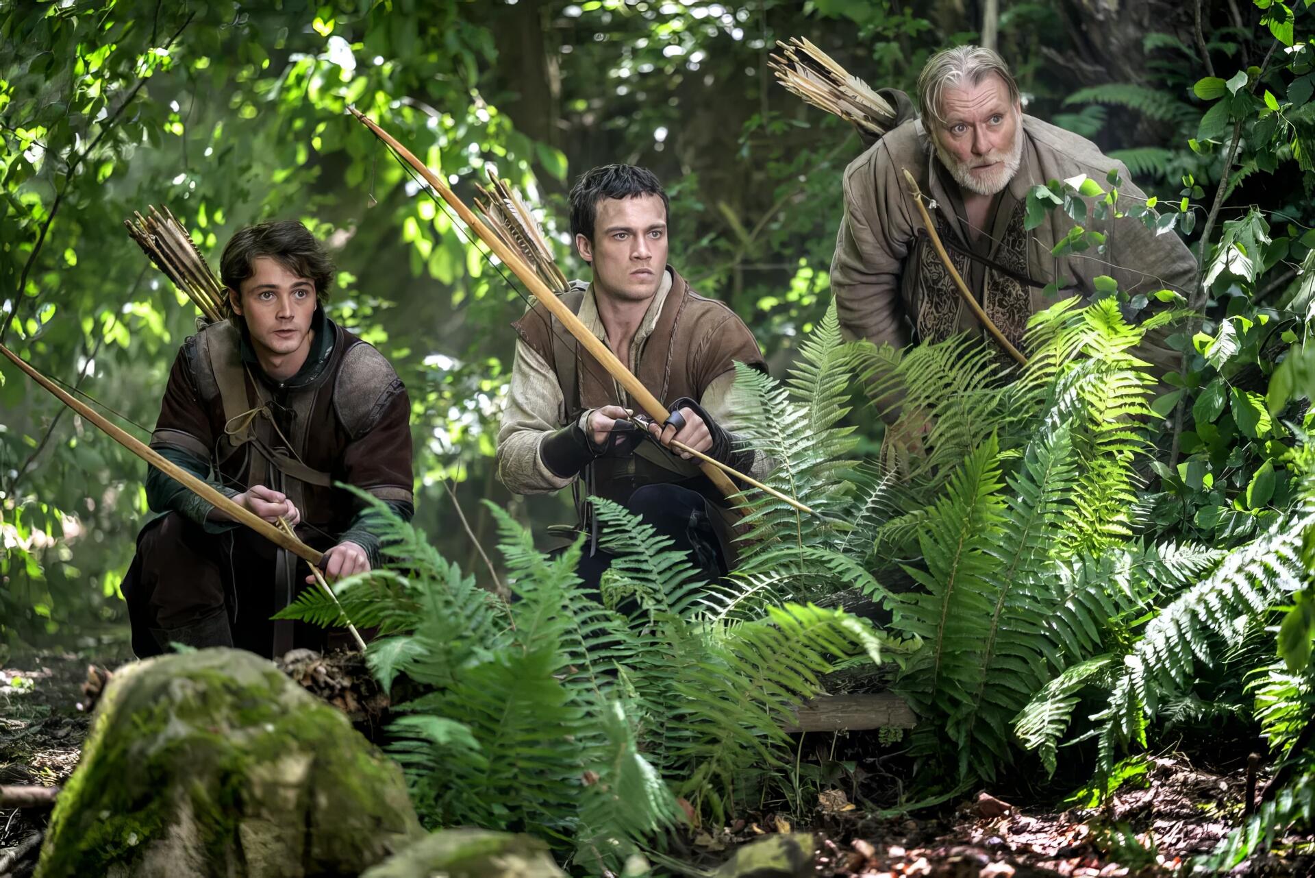 HD PC desktop wallpaper: three archers crouch amid ferns in a dense forest, scene from the TV show Robin Hood (2025).