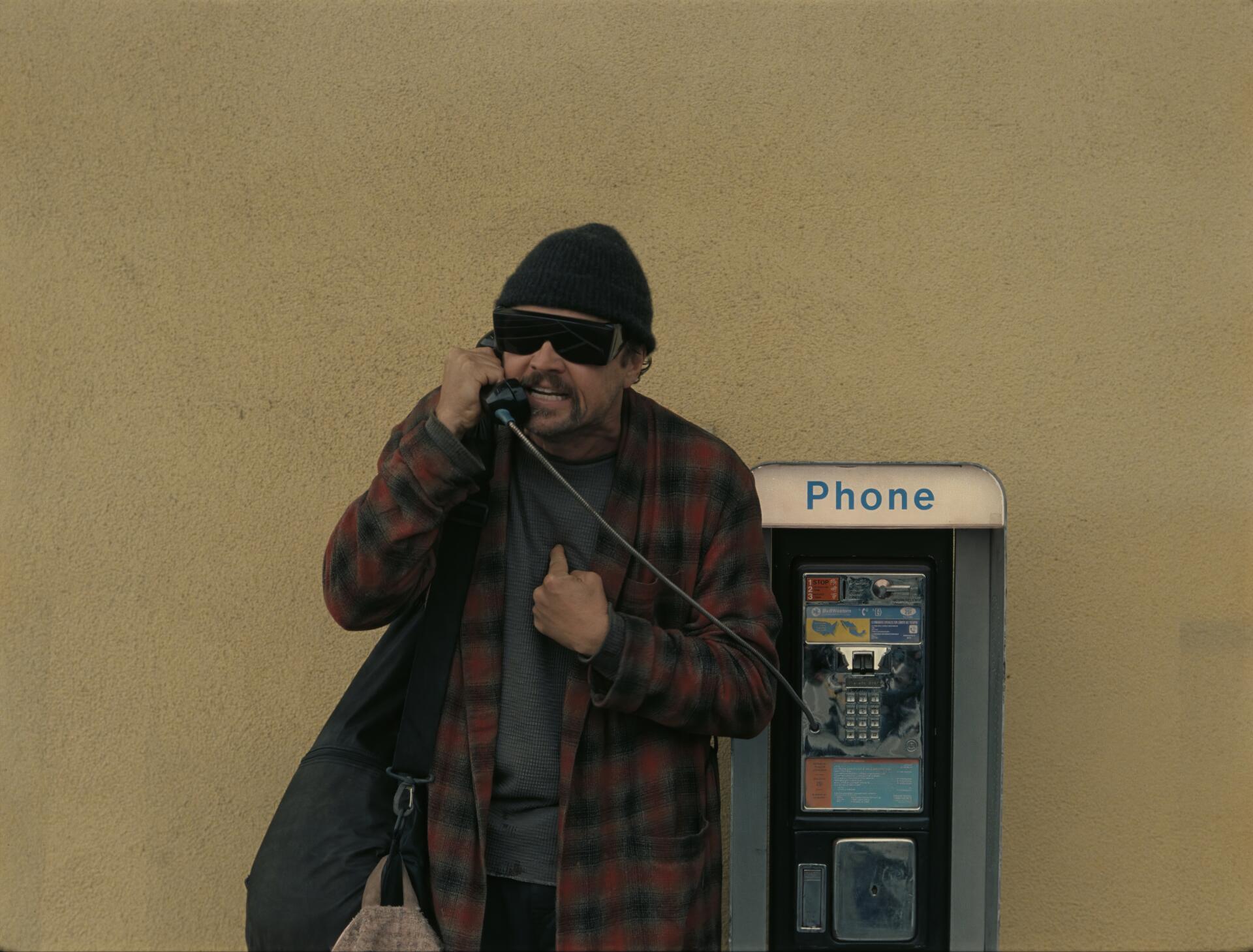 4K Ultra HD PC wallpaper: still from the movie One Battle After Another — a bearded man in a beanie and sunglasses using a payphone against a beige wall.