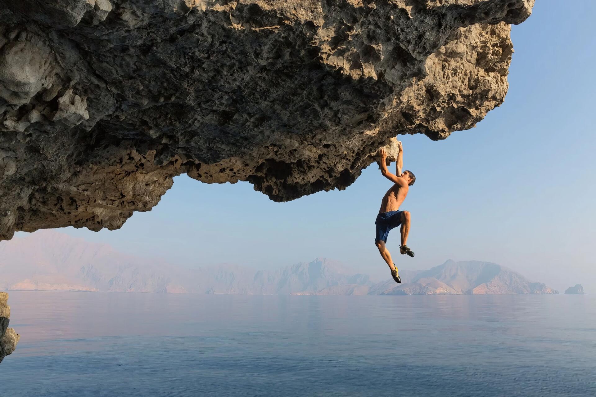Sport climber hangs from an overhanging cliff above a tranquil sea; identity not specified. 2K Quad HD PC desktop wallpaper.