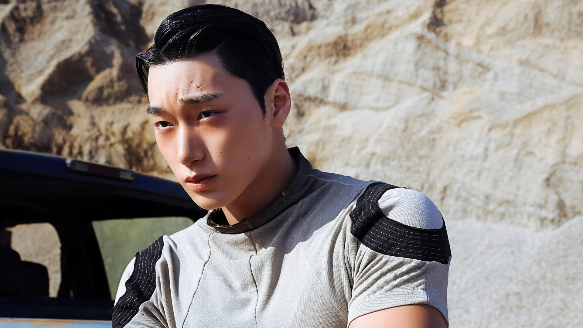 4K K-pop desktop wallpaper inspired by ATEEZ: intense young man in a futuristic outfit with a forehead scar, posed against sunlit rocky desert background.