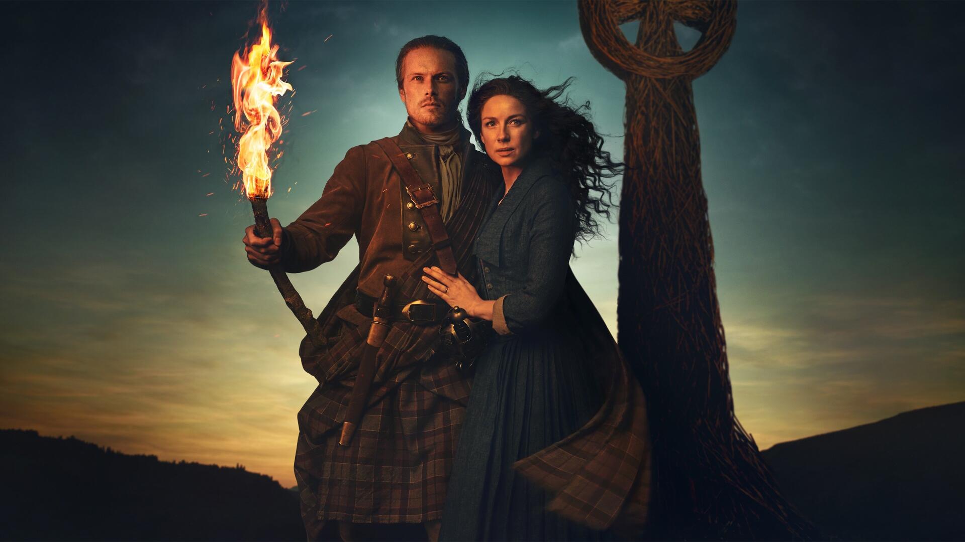 Outlander 4K Ultra HD PC desktop wallpaper: a man in a kilt holding a torch and a woman in period dress embrace beside a standing Celtic cross at sunset.