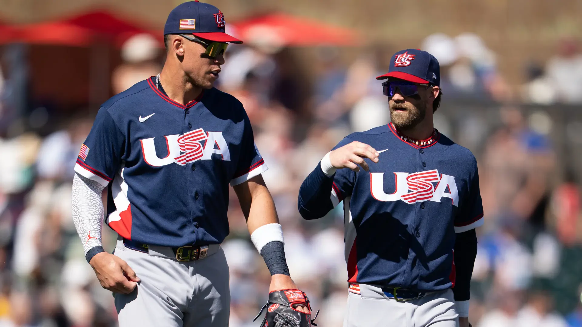 HD PC desktop wallpaper: two USA Baseball players in navy USA uniforms walking and talking across a sunlit stadium field.