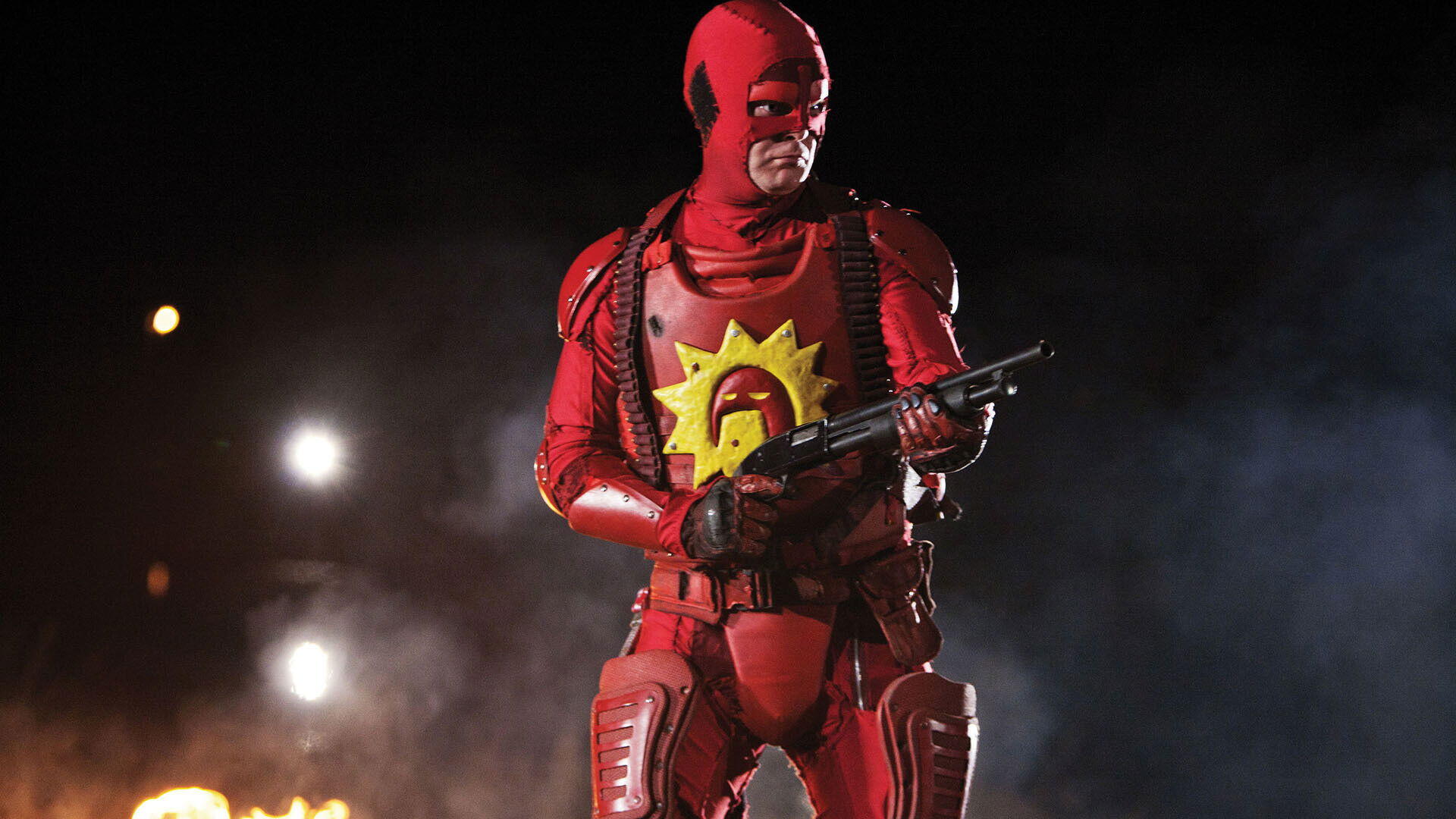 HD PC desktop wallpaper background of a red-armored vigilante with a yellow sun emblem on his chest, holding a shotgun amid smoke, sparks and distant flames.