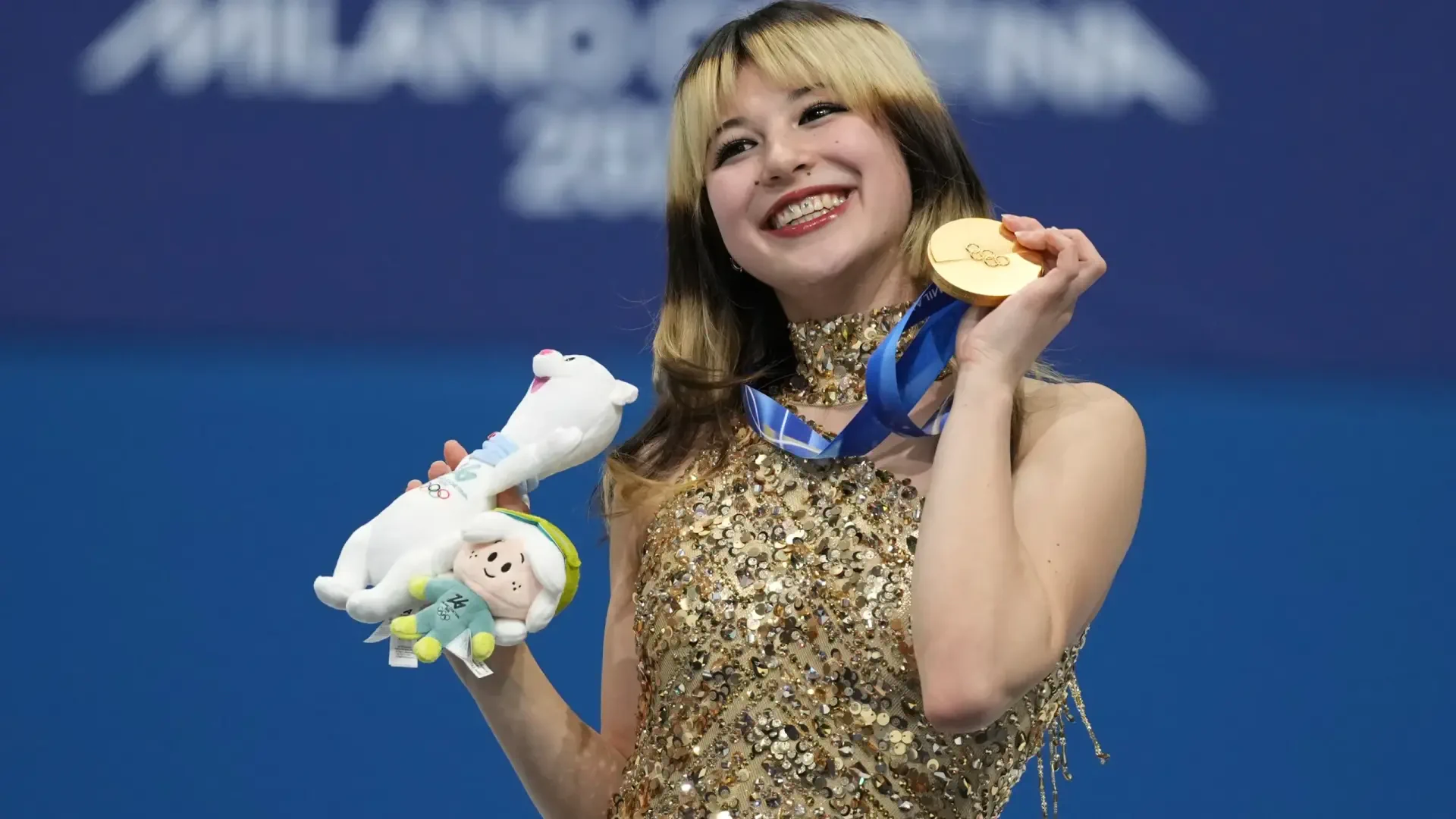 Smiling female figure skater holding a gold medal and plush mascot at the Winter Olympics, ice skating sports scene rendered as a 2K Quad HD desktop wallpaper.