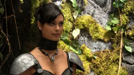 Orici from Stargate SG-1 in leather armor and a choker, posed before mossy rocks and foliage — cinematic 5K Ultra HD PC desktop wallpaper.