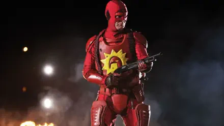 HD PC desktop wallpaper background of a red-armored vigilante with a yellow sun emblem on his chest, holding a shotgun amid smoke, sparks and distant flames.