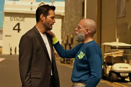 Two actors on a studio backlot in conversation — one in a dark blazer, the other in a blue sweatshirt with a beard — promotional image for the film Outcome (2026). 2K Quad HD desktop wallpaper.