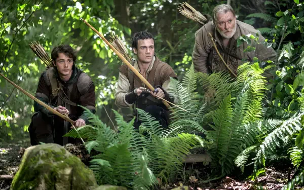 HD PC desktop wallpaper: three archers crouch amid ferns in a dense forest, scene from the TV show Robin Hood (2025).