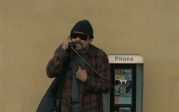 4K Ultra HD PC wallpaper: still from the movie One Battle After Another — a bearded man in a beanie and sunglasses using a payphone against a beige wall.
