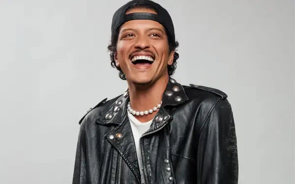 HD PC desktop wallpaper of a smiling music celebrity in a black leather jacket, backward cap and pearl necklace, posed against a light gray background.