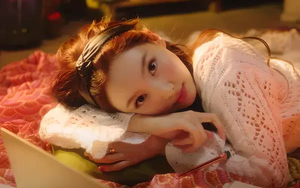 2K Quad HD PC wallpaper of a cute K-pop idol reclining on a bed, head on folded arms, wearing a knit sweater and headband, bathed in warm, cozy lighting.