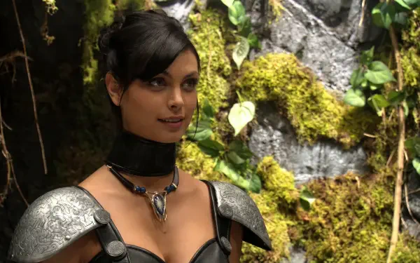 Orici from Stargate SG-1 in leather armor and a choker, posed before mossy rocks and foliage — cinematic 5K Ultra HD PC desktop wallpaper.