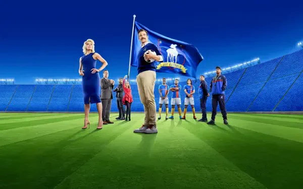 Ted Lasso 4K Ultra HD PC desktop wallpaper: the coach stands center holding a soccer ball, flanked by teammates and staff on a brightly lit stadium pitch under night lights.