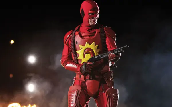 HD PC desktop wallpaper background of a red-armored vigilante with a yellow sun emblem on his chest, holding a shotgun amid smoke, sparks and distant flames.