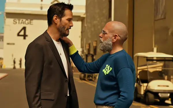 Two actors on a studio backlot in conversation — one in a dark blazer, the other in a blue sweatshirt with a beard — promotional image for the film Outcome (2026). 2K Quad HD desktop wallpaper.