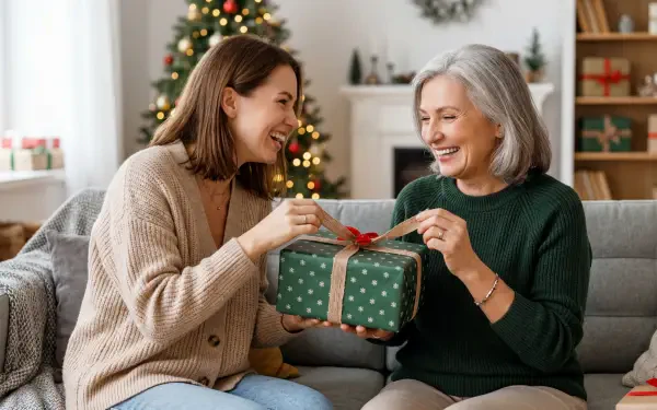 Gift giving — two smiling women exchange a wrapped green present on a couch in a cozy living room with a decorated Christmas tree; 5K Ultra HD PC desktop wallpaper and background.