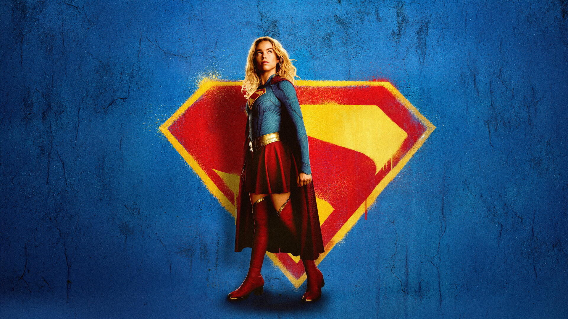 Supergirl (2026) stands in a heroic pose before a large S emblem on a blue textured wall — 4K Ultra HD PC desktop wallpaper.