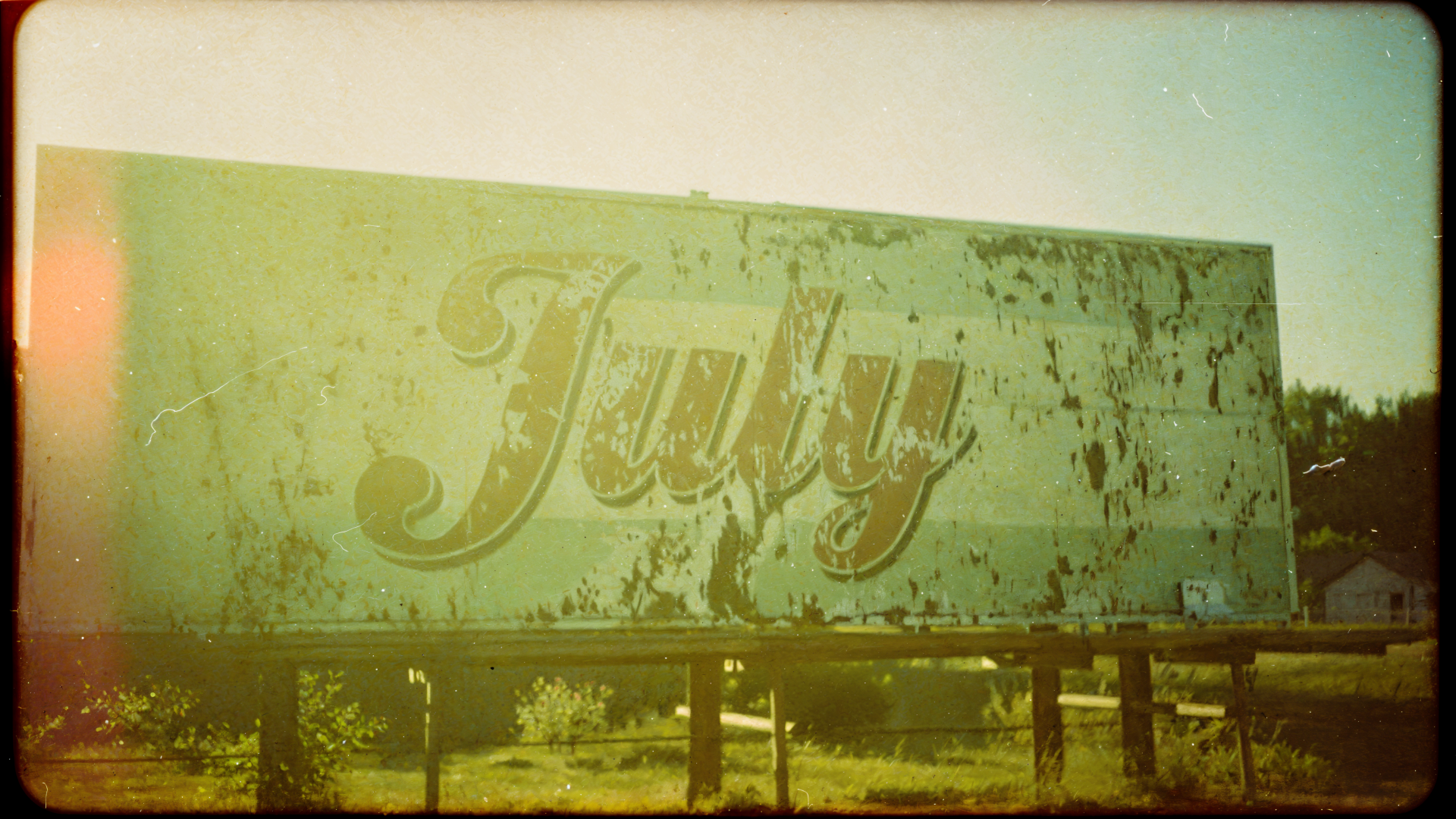 Weathered billboard with cursive July amid sunlit, faded green-yellow tones — 4K Ultra HD PC desktop wallpaper and background.