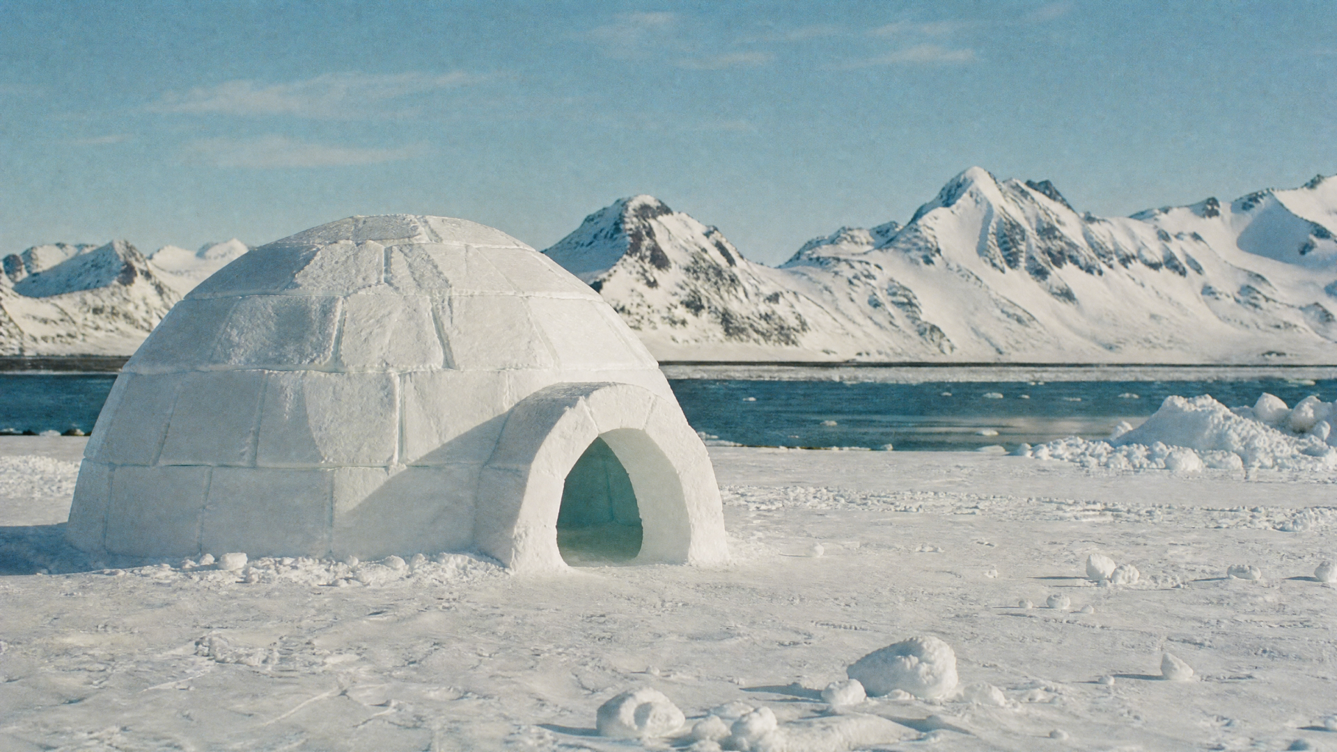 Igloo on a frozen shoreline with distant snow-capped mountains and clear blue sky — 4K Ultra HD PC desktop wallpaper and background.