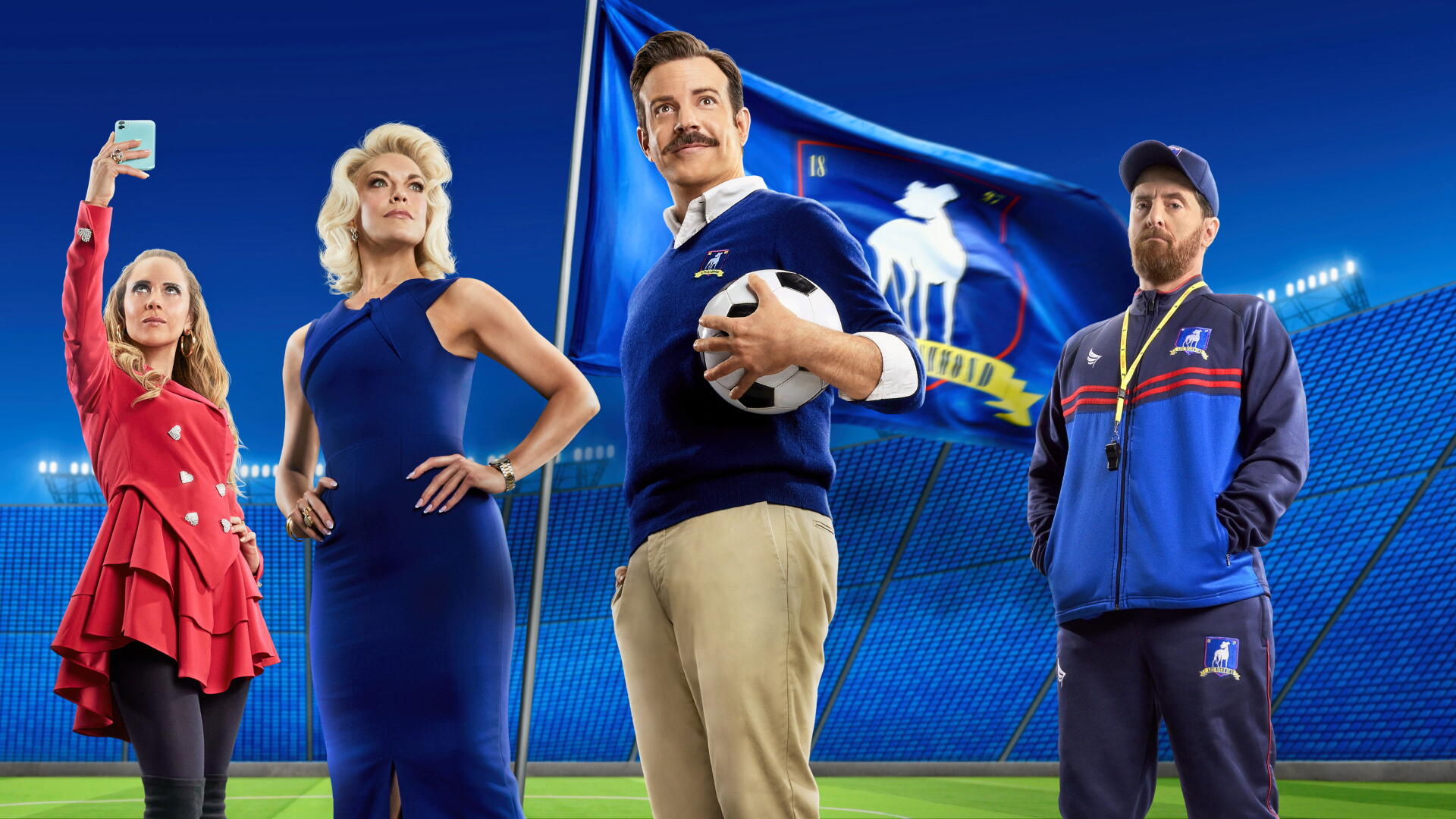 Ted Lasso cast posing in a stadium, central coach with a mustache holding a soccer ball against vivid blue banners — 4K Ultra HD PC desktop wallpaper and background.