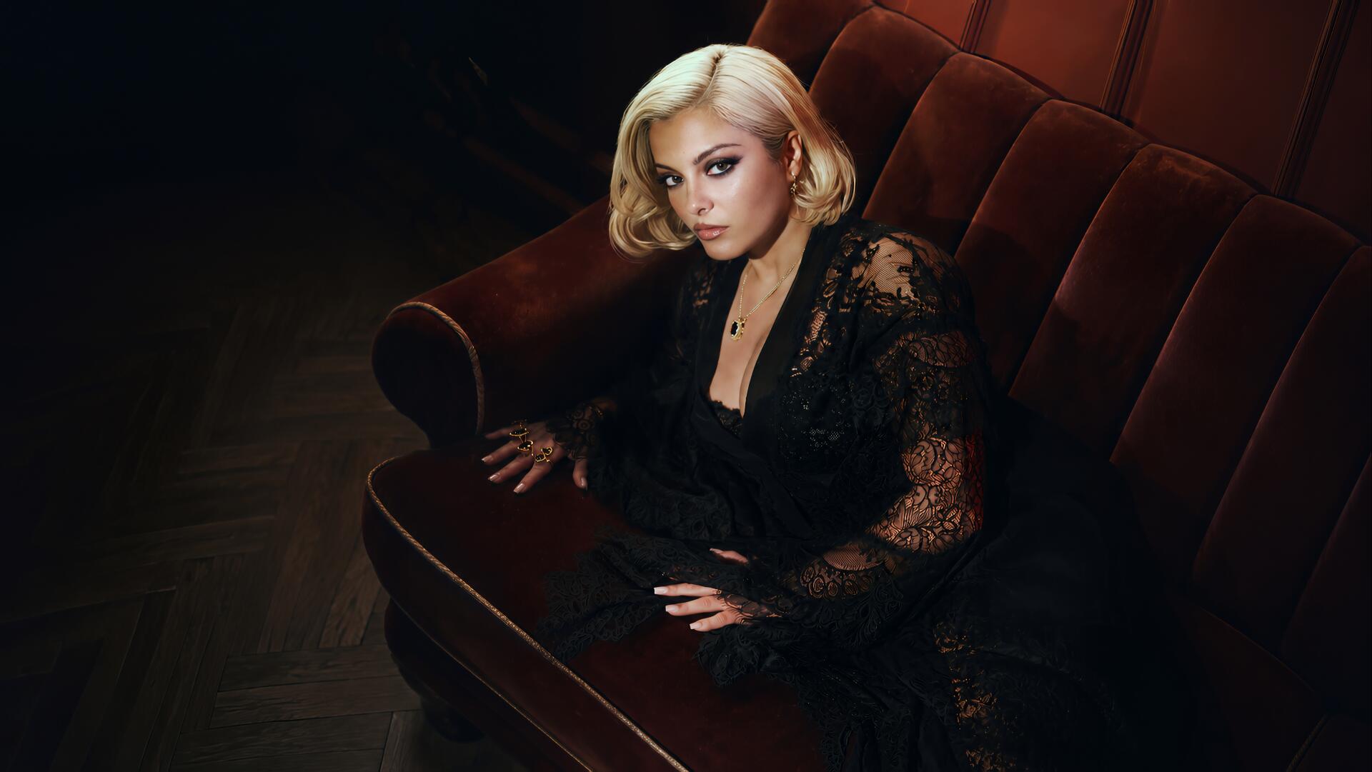 Celebrity singer seated on a velvet sofa in moody lighting, blonde hair and black lace outfit — 4K Ultra HD PC desktop wallpaper/background.