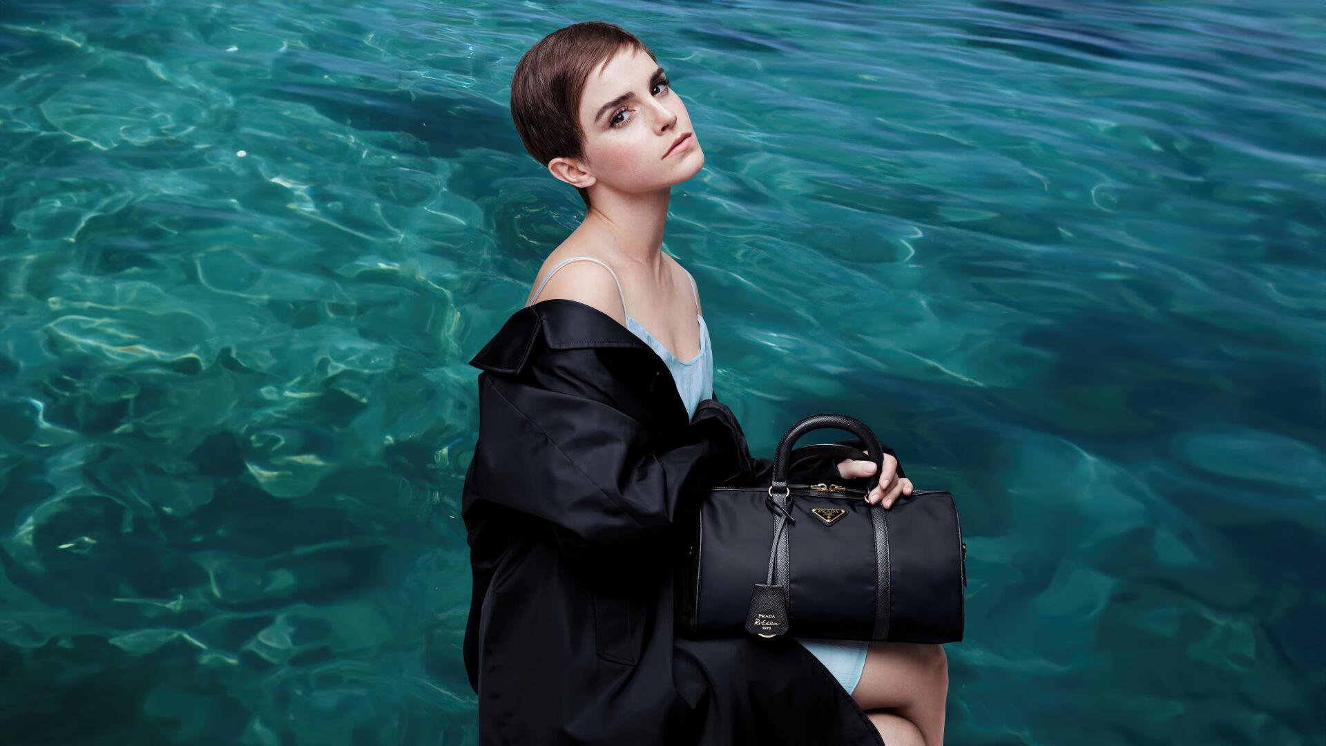 4K Ultra HD desktop wallpaper: short-haired woman in a black coat seated by clear turquoise water, gazing upward while holding a structured black handbag.