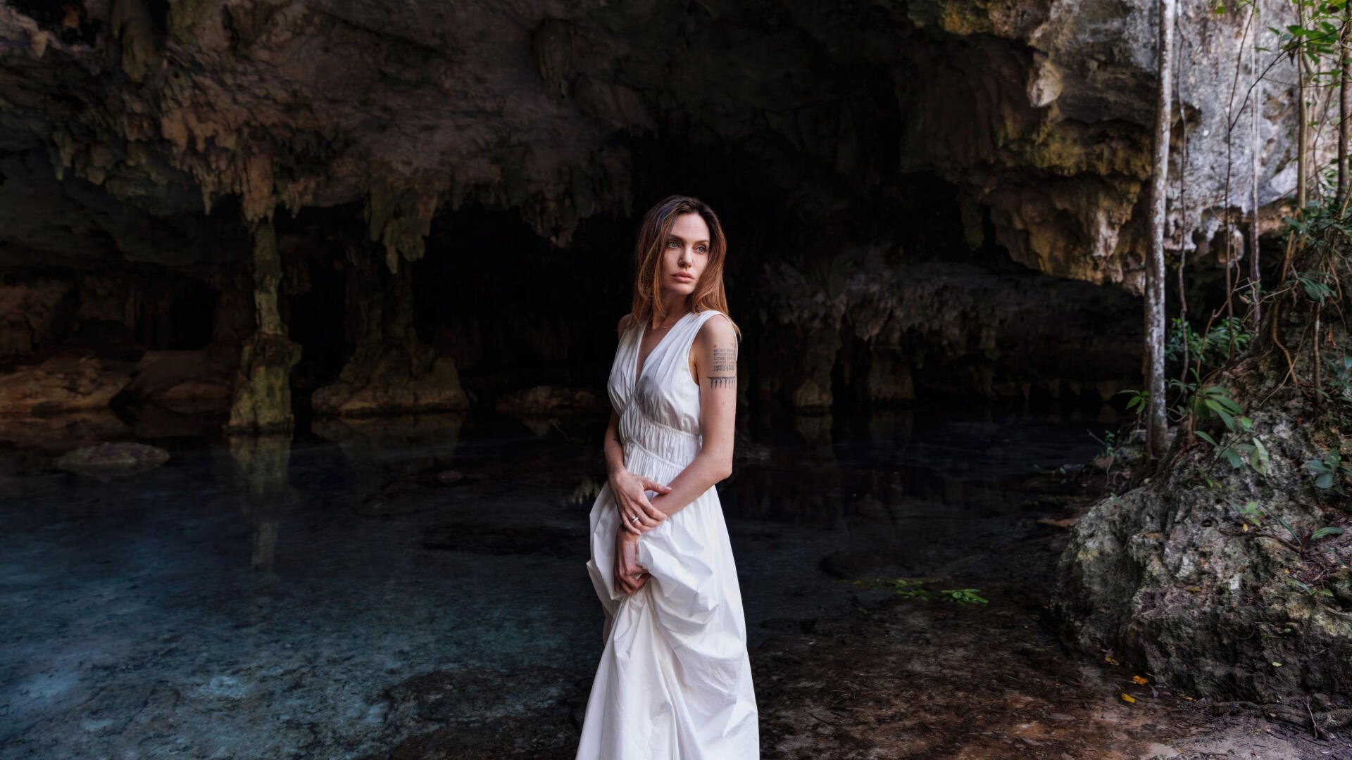 Celebrity actress in a flowing white gown standing at a cave's edge over clear water, moody 4K Ultra HD PC desktop wallpaper/background.