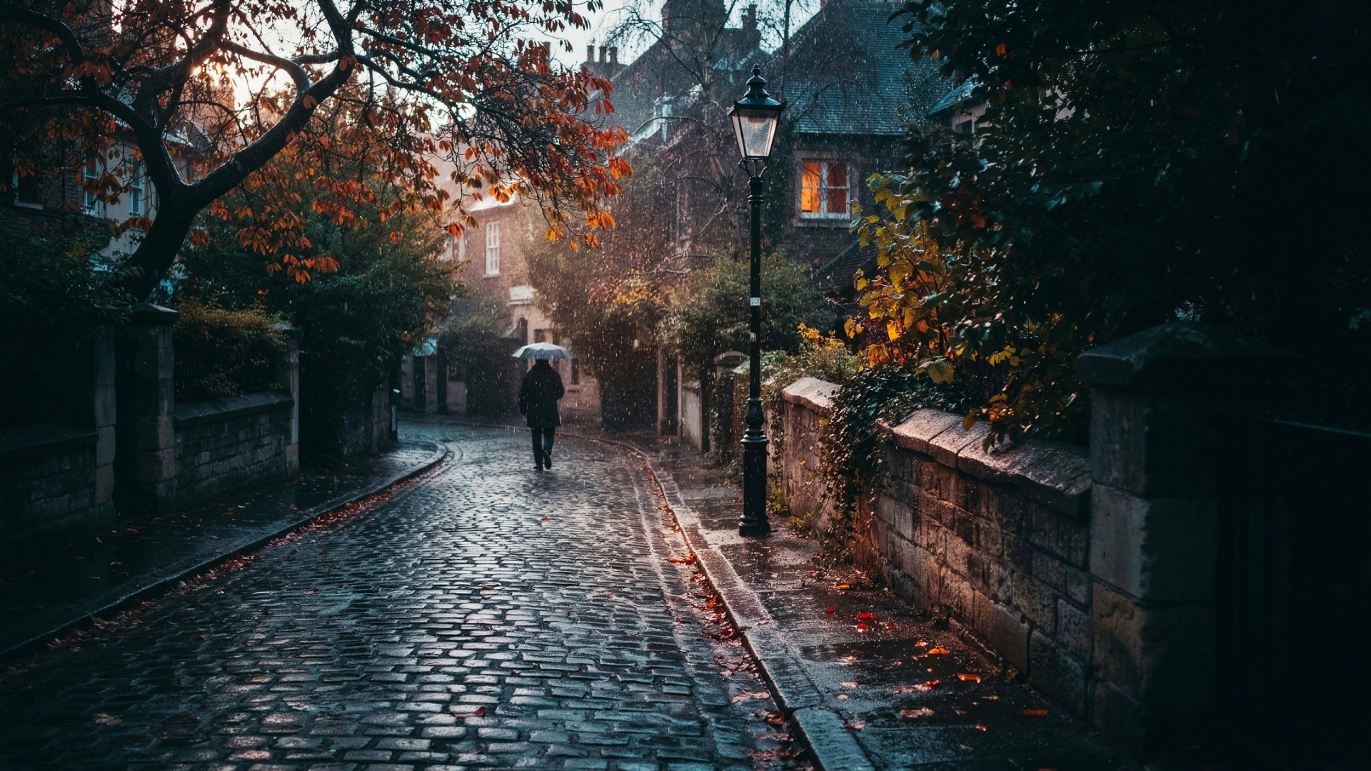 Rainy fall cobblestone alley street at dusk with a lone figure under an umbrella; warm lamp light and wet reflections — 2K Quad HD PC desktop wallpaper/background.