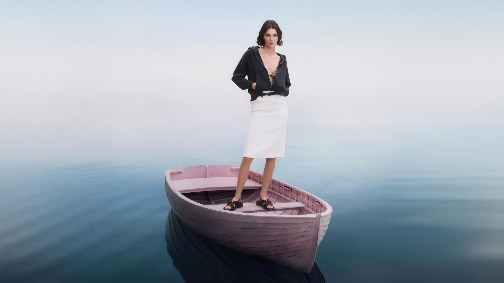 Model stands on a small wooden boat drifting on glassy water, wearing a black jacket and white skirt — 4K Ultra HD PC desktop wallpaper.