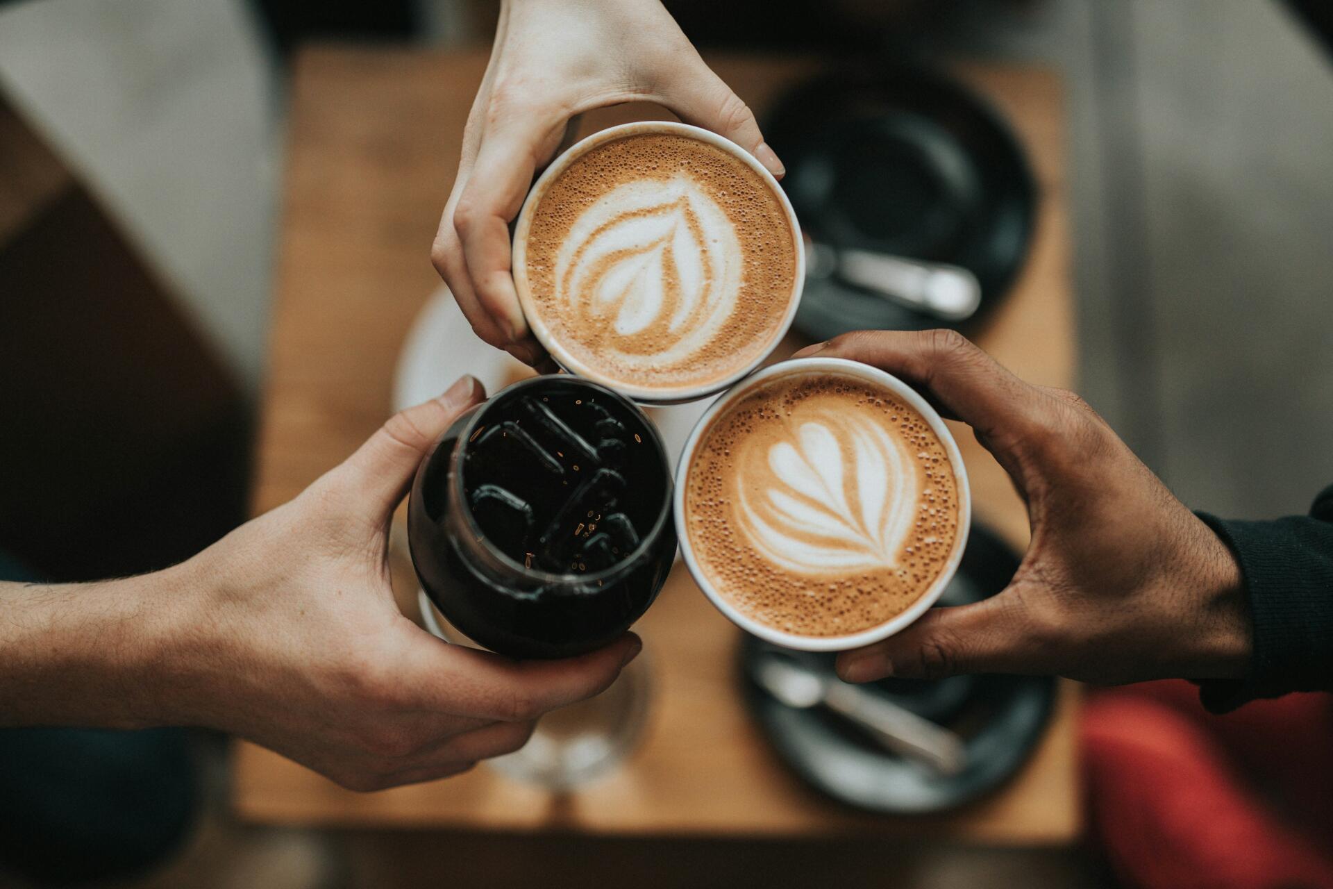 Espresso Fall cozy scene: three hands raise two lattes with leaf art and an iced espresso, warm autumn tones — 4K Ultra HD PC desktop wallpaper and background.