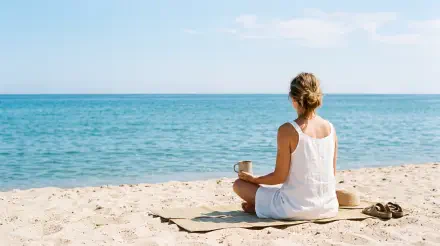 Minimalist summer 5K Ultra HD PC desktop wallpaper: person in white dress sits on sandy beach, holding a mug and gazing at calm turquoise sea under a pale blue sky.