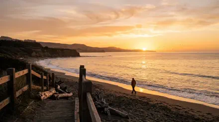 Golden sunrise over a quiet beach with a lone walker along the shore; cliff silhouettes and wooden railing in foreground — 5K Ultra HD PC desktop wallpaper and background.
