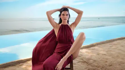 Model in a flowing burgundy halter dress seated by an infinity pool on a sandy beach — 4K Ultra HD PC desktop wallpaper/background.