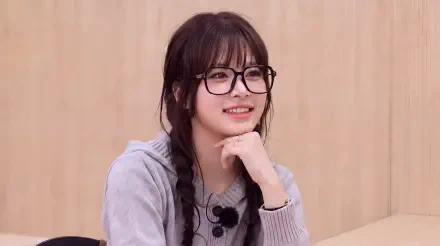 Smiling K-pop idol in glasses with braided hair and a gray sweater, seated in a light studio — 5K Ultra HD PC desktop wallpaper/background.