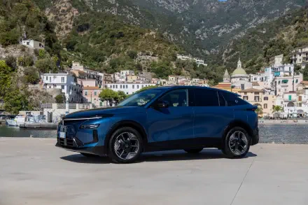 Blue Lancia Gamma electric SUV parked by a waterfront village and mountain backdrop, HD desktop wallpaper.
