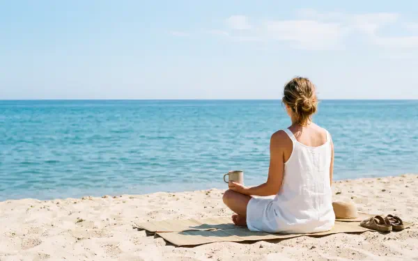 Minimalist summer 5K Ultra HD PC desktop wallpaper: person in white dress sits on sandy beach, holding a mug and gazing at calm turquoise sea under a pale blue sky.