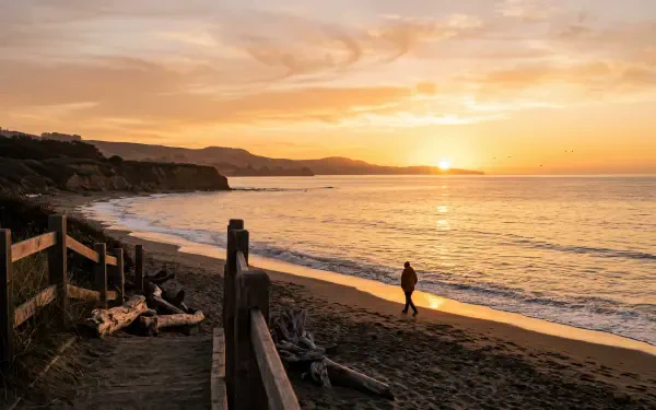 Golden sunrise over a quiet beach with a lone walker along the shore; cliff silhouettes and wooden railing in foreground — 5K Ultra HD PC desktop wallpaper and background.