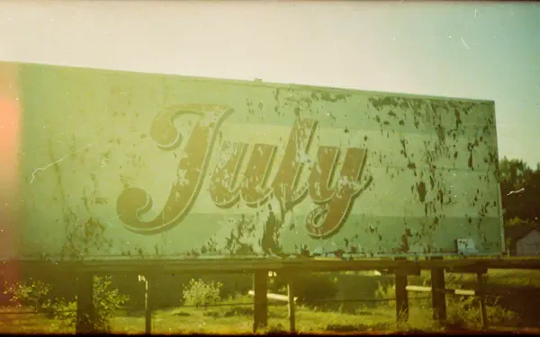 Weathered billboard with cursive July amid sunlit, faded green-yellow tones — 4K Ultra HD PC desktop wallpaper and background.