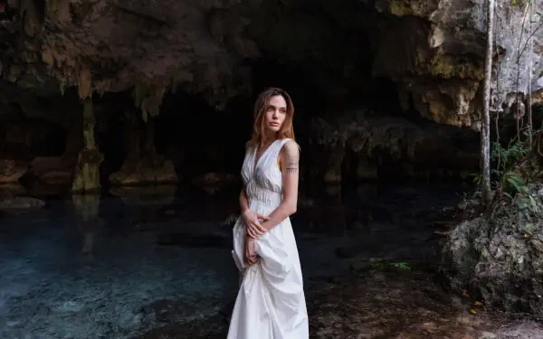 Celebrity actress in a flowing white gown standing at a cave's edge over clear water, moody 4K Ultra HD PC desktop wallpaper/background.