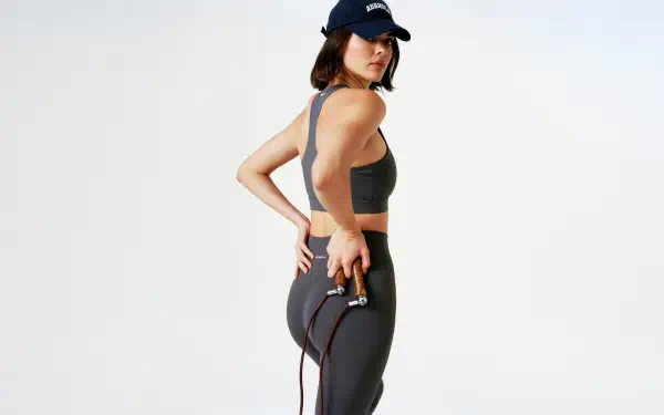 5K Ultra HD PC wallpaper of a woman in dark athletic wear and cap, side pose holding a jump rope against a clean, light background.