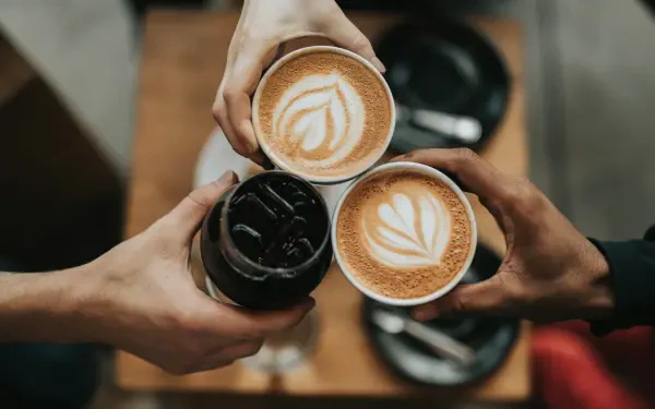 Espresso Fall cozy scene: three hands raise two lattes with leaf art and an iced espresso, warm autumn tones — 4K Ultra HD PC desktop wallpaper and background.